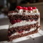 A decadent slice of Gothic Black Forest Cake with layers of chocolate cake, whipped cream, and cherries, topped with fresh cherries.