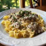 A close-up of a plate piled high with egg noodles topped with creamy ground beef stroganoff and garnished with fresh parsley.