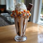 A tall Halloween Candy Milkshake topped with whipped cream, candy corn, chocolate, and a candy bar.
