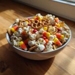 A close-up of a white bowl filled with a festive Halloween Snack Mix, featuring popcorn, pretzels, candy corn, and M&Ms.