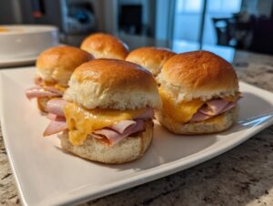 Close-up of several delicious ham and cheese sliders on a white plate, with melted cheese oozing out.