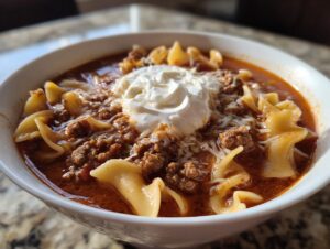 A close-up of a bowl of hearty Lasagna Soup, featuring noodles, seasoned ground meat, and topped with a dollop of sour cream and shredded cheese.