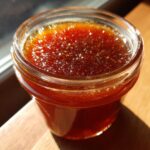 Close-up of a glass jar filled with rich, dark apple butter, glistening in the sunlight.