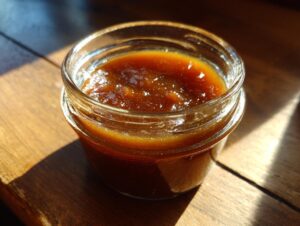 A close-up of a small glass jar filled with rich, dark apple butter, bathed in sunlight.