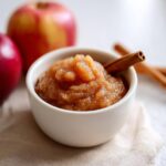 A close-up of a bowl filled with textured homemade applesauce, garnished with a cinnamon stick. Fresh apples and more cinnamon sticks are blurred in the background.