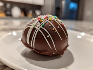 A close-up of a dark chocolate hot chocolate bomb topped with white drizzle and colorful sprinkles on a white plate.