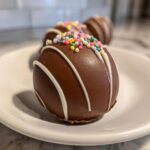 Close-up of a festive hot chocolate bomb topped with colorful sprinkles and white chocolate drizzle on a white plate.