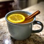 A close-up of a steaming Hot Toddy in a blue mug, garnished with a lemon slice and cinnamon sticks.