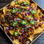 A close-up view of a baking sheet filled with loaded nachos, topped with seasoned ground beef, melted cheese, red onions, and jalapeño slices.