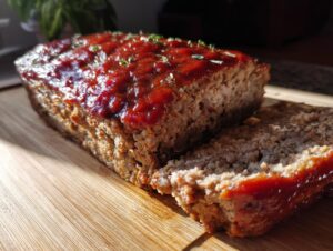A close-up of a moist meatloaf topped with a glossy glaze and fresh herbs, with a slice cut and resting beside it on a wooden cutting board.
