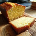 A close-up of a moist pound cake, with one slice cut and placed in front of the loaf on a wooden board.