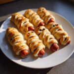 A plate of golden-brown Mummy Hot Dogs, wrapped in pastry with olive eyes, ready for Halloween.