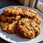 A close-up of several golden-brown oatmeal cookies with visible raisins, piled on a white plate.