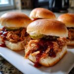 Close-up of four savory pulled pork sliders on a white plate, topped with pickles and BBQ sauce.