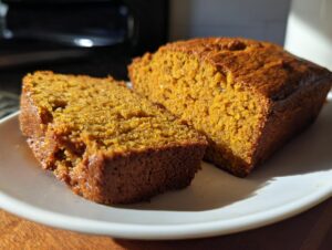 A slice of moist Pumpkin Beer Bread sits next to the loaf on a white plate, bathed in warm sunlight.