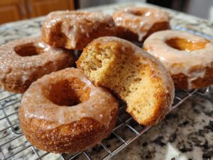 A close-up of freshly baked Pumpkin Donuts on a cooling rack, some with a glaze and others with cinnamon sugar. One donut is broken in half to show its texture.