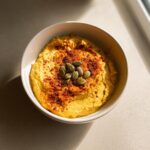 A bowl of vibrant Pumpkin Hummus topped with pumpkin seeds and paprika.