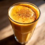 A close-up of a vibrant orange pumpkin smoothie in a glass, topped with a sprinkle of cinnamon.