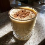 A close-up of rich and creamy eggnog in a decorative glass, topped with a sprinkle of cinnamon.