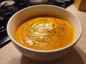 A close-up of a white bowl filled with vibrant orange Roasted Butternut Squash Soup, swirled with cream and sprinkled with herbs.