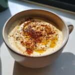 A close-up of a bowl of velvety roasted cauliflower soup, garnished with paprika, black pepper, and a drizzle of olive oil.