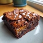 Close-up of a decadent salted caramel brownie slice, showing gooey caramel filling and flaky sea salt on top.