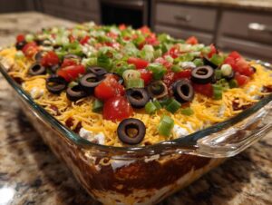 A close-up view of a freshly prepared Seven Layer Dip in a clear glass dish, topped with cheese, olives, tomatoes, and green onions.