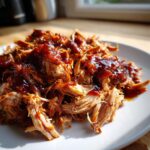 A close-up of tender, shredded slow cooker pulled pork coated in glistening BBQ sauce on a white plate.