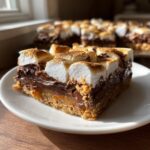 A close-up of a decadent S'mores Bar on a white plate, featuring a graham cracker crust, gooey chocolate, caramel, and perfectly toasted marshmallows.