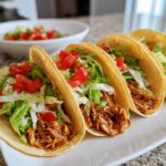 A close-up of three Soft Chicken Tacos filled with shredded chicken, lettuce, and diced tomatoes on a white plate.