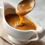 A close-up of rich, smooth turkey gravy being spooned from a metal spoon into a white gravy boat.