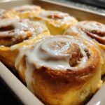 Close-up of freshly baked cinnamon rolls covered in creamy white glaze in a baking dish.