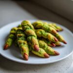 A pile of green Witch Finger Cookies with almond fingernails and red tips on a white plate.