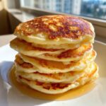 A tall stack of golden brown, fluffy pancakes drizzled with syrup, ready to eat.