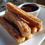 A stack of freshly fried, golden brown churros heavily coated in cinnamon sugar, served with chocolate dipping sauce.