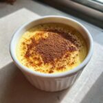 A creamy yellow egg custard served in a white ramekin, topped with a dusting of cocoa powder.
