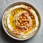 Overhead view of creamy hummus in a bowl, drizzled with olive oil, chili flakes, and whole chickpeas.