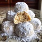 A stack of powdered sugar-dusted russian tea cakes, with one cookie broken open revealing the crumbly texture inside.