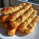 A close-up stack of soft, golden garlic breadsticks topped with melted cheese and herbs.