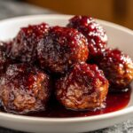 A close-up shot of several glistening Grape Jelly Meatballs coated in a thick, dark red sauce, served in a white bowl.
