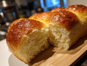 Close-up of fluffy, golden-brown baked dinner rolls on a wooden board, one roll split open.