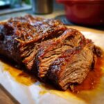 Close-up of tender, juicy braised brisket, sliced and glistening with sauce on parchment paper.