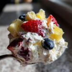 Close-up of a spoonful of creamy cheesecake fruit salad mixed with strawberries, blueberries, and pineapple chunks.