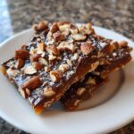 Close-up of rich chocolate almond toffee pieces on a white plate, showcasing the crunchy nuts and glossy toffee.