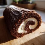 A close-up of a chocolate Yule log cake rolled with white cream filling, dusted with powdered sugar.
