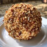 A perfectly formed classic cheese ball coated in chopped pecans, presented on a white plate.