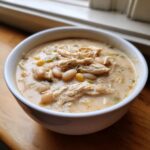 A close-up of a white bowl filled with creamy chicken chili, featuring shredded chicken, white beans, and corn.