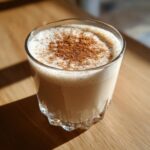 A close-up of a glass filled with creamy Coquito, topped with a dusting of cinnamon.