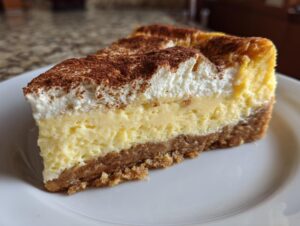 A thick slice of Eggnog cheesecake showing graham cracker crust, creamy filling, and whipped topping dusted with cocoa.