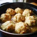Close-up of several fluffy, golden-brown dumplings simmering in a savory sauce inside a dark skillet.